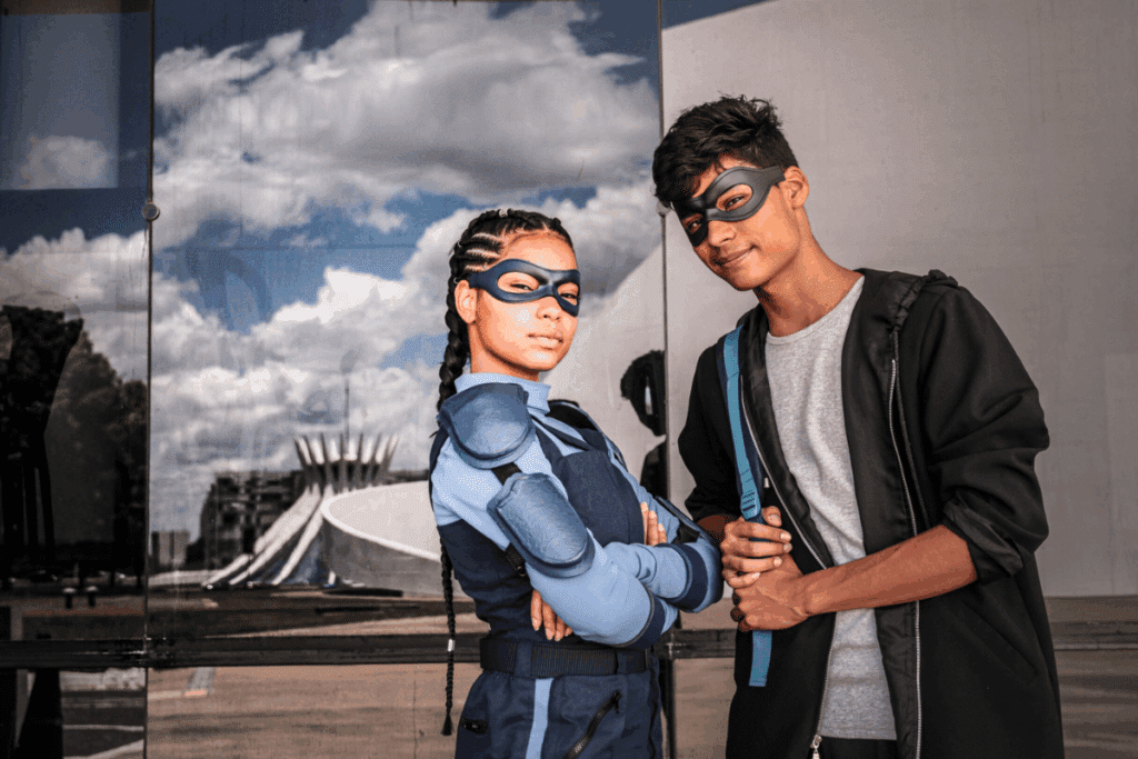Fotografia de dois jovens posando lado a lado usando máscaras pretas nos olhos. À esquerda, uma garota com tranças nagô veste um traje tático azul com protetores nos ombros. À direita, um garoto de jaqueta preta segura as alças de uma mochila. Ao fundo, uma parede espelhada reflete a Catedral de Brasília e o céu com nuvens.