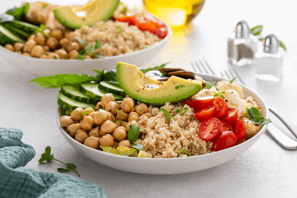 Prato fundo com salada composta por grão-de-bico, quinoa, tomates-cereja cortados ao meio, rodelas de pepino e uma fatia de abacate no topo, sobre uma superfície clara ao lado de galheteiros de sal e pimenta.