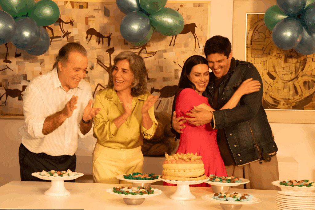Quatro pessoas comemorando ao redor de uma mesa decorada com bolo, doces e balões azuis e verdes; um homem e uma mulher mais velha aplaudem sorrindo enquanto um casal jovem se abraça.