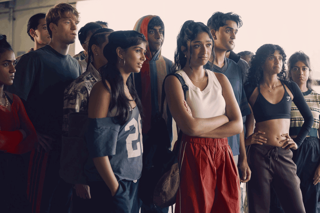 Grupo de jovens reunidos, com diferentes pessoas vestindo roupas casuais e modernas. Eles aparecem lado a lado em um ambiente amplo, com uma jovem de regata branca e calça vermelha em destaque no centro.