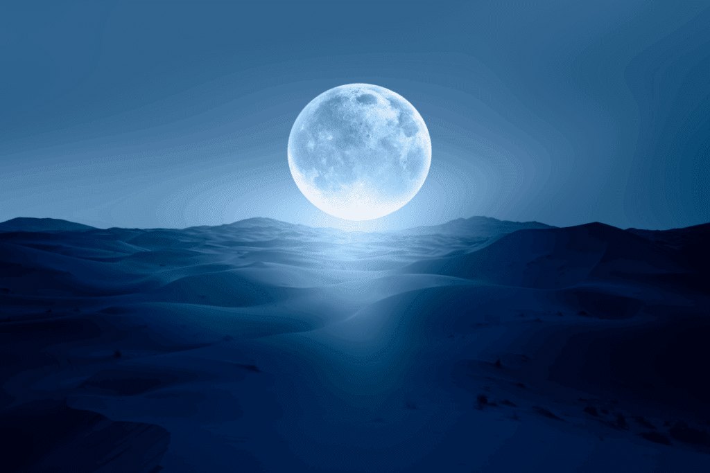A imagem mostra uma Lua Cheia brilhando sobre uma paisagem de dunas, iluminando o cenário noturno em tons de azul. A lua aparece em destaque no horizonte, enquanto as dunas se estendem ao fundo