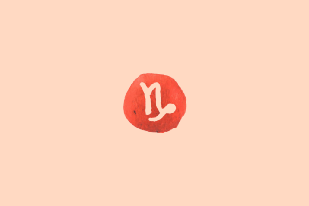Ilustração do signo de Capricórnio sobre fundo uniforme em tom pêssego-claro. No centro, há uma mancha circular irregular em vermelho-coral, com textura de aquarela e pequenas variações de tonalidade. O símbolo de Capricórnio aparece em tom creme, composto por traços curvos e alongados que formam uma figura semelhante à letra M, com uma das extremidades prolongada em uma curva arredondada voltada para baixo.