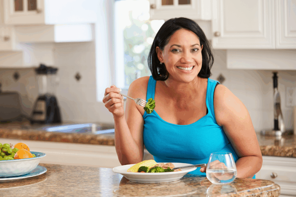 Mulher sentada à mesa em uma cozinha, sorrindo enquanto segura um garfo com um pedaço de brócolis. Ela tem o cabelo curto preto e veste uma regata azul e tem um prato com vegetais à sua frente, além de uma tigela com frutas ao lado.