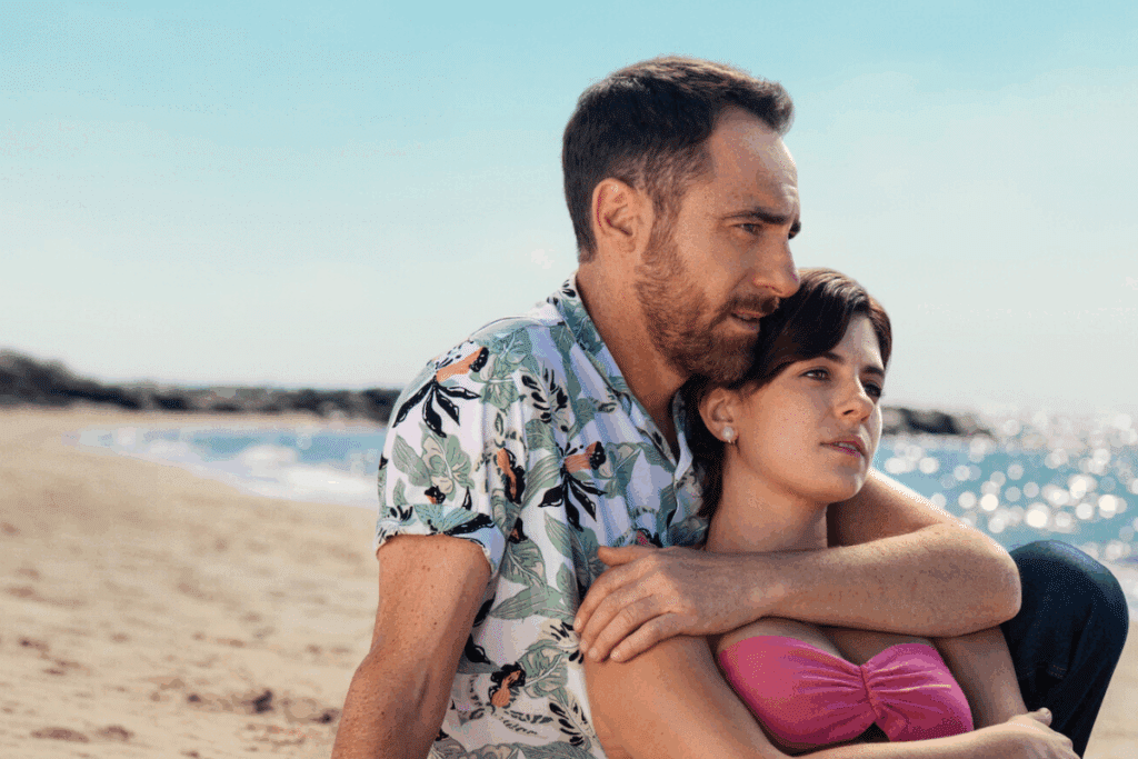Homem abraçando uma mulher por trás, sentados em uma praia ensolarada.