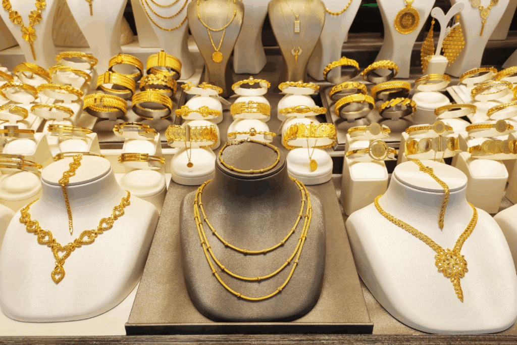 Vitrine de joalheria repleta de joias de ouro exibidas em suportes brancos e pretos, incluindo colares elaborados, braceletes, pulseiras e brincos pendentes.