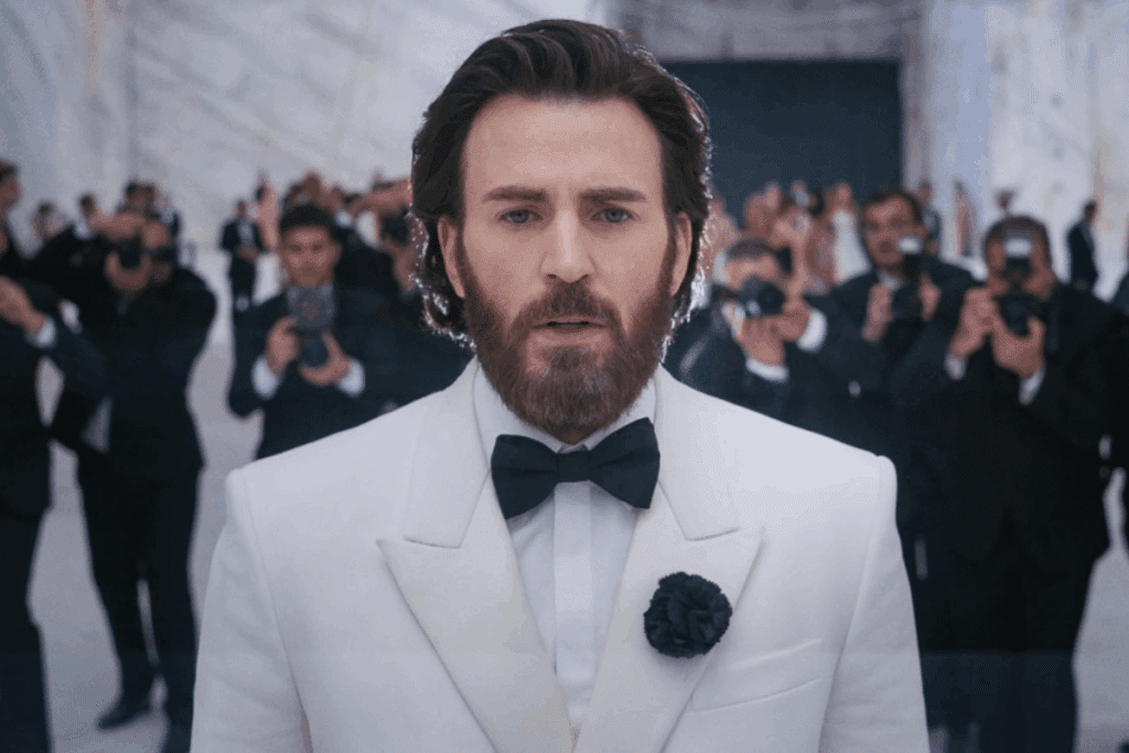 Um homem de barba vestindo um terno elegante com paletó branco, gravata borboleta preta e uma flor preta na lapela olha diretamente para a frente. Ao fundo, uma multidão de fotógrafos e fotógrafas de roupa preta segura câmeras apontadas na direção dele.