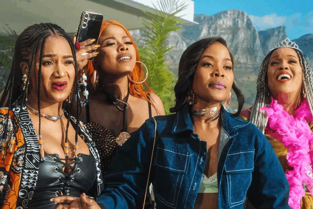 Quatro mulheres negras vestidas em trajes elegantes e modernos expressam diferentes reações ao ar livre, com montanhas ao fundo.