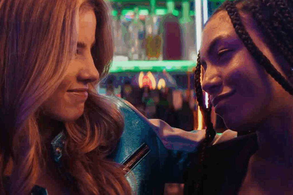 Giulia Be e Any Gabrielly em “As Dez Vantagens de Morrer Depois de Você”: duas amigas se olham sorrindo, abraçadas, em um bar iluminado por luzes de neon coloridas.