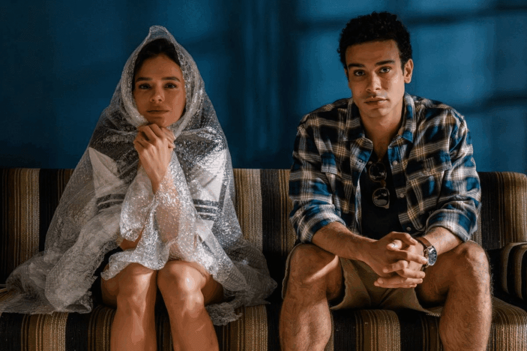 Um casal está sentado em um sofá. A mulher usa uma camiseta branca de mangas curtas e uma saia curta escura, cobertas por um plástico bolha, que também envolve sua cabeça. O homem veste uma camiseta preta, uma camisa xadrez em tons de azul e branco aberta sobre ela e um short bege. Ele também usa um relógio de pulso e óculos escuros pendurados na gola da camiseta.